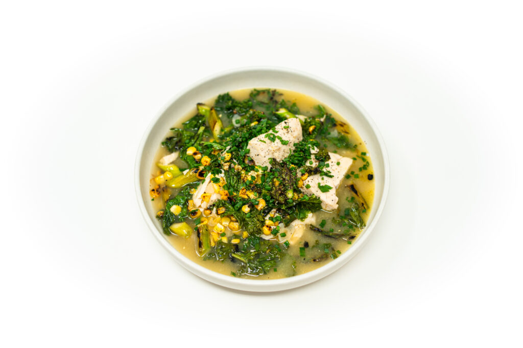 Flaky corn poached amberjack served in a rich sweet corn broth with blistered kale, charred asparagus, and fresh chives in a shallow white bowl.