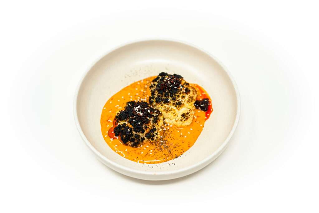 Heavily charred cauliflower florets resting in a vibrant orange fermented chili emulsion, topped with chili crisp and toasted sesame seeds.