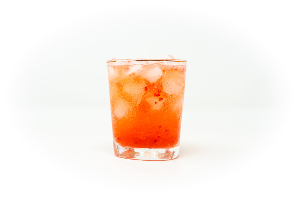 A bright red spicy strawberry rosé spritz poured over ice in a thick rocks glass, backed by bottles of Alma Tepec, triple sec, and brut wine.