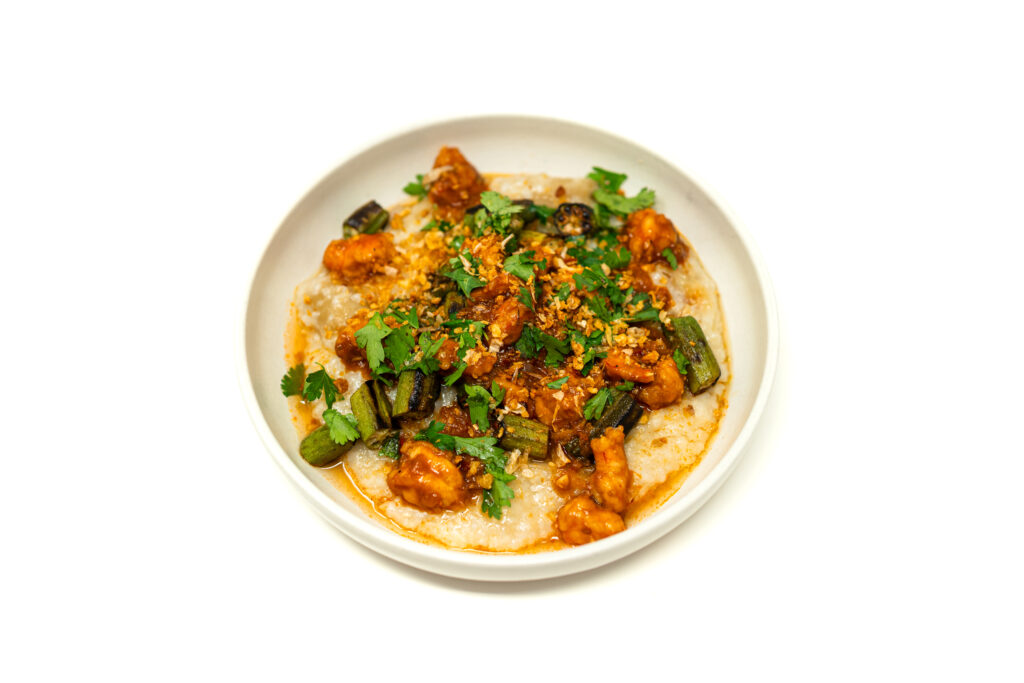 Heavy overhead lighting casting shadows across a shallow bowl of creamy jasmine rice porridge, topped with aggressively charred okra and glossy red chili shrimp.