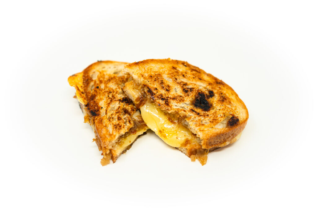 Overhead shot of a halved grilled cheese sandwich on a wooden board, pulling melted white cheddar across dark fig and onion jam with crispy shallots catching harsh natural light.