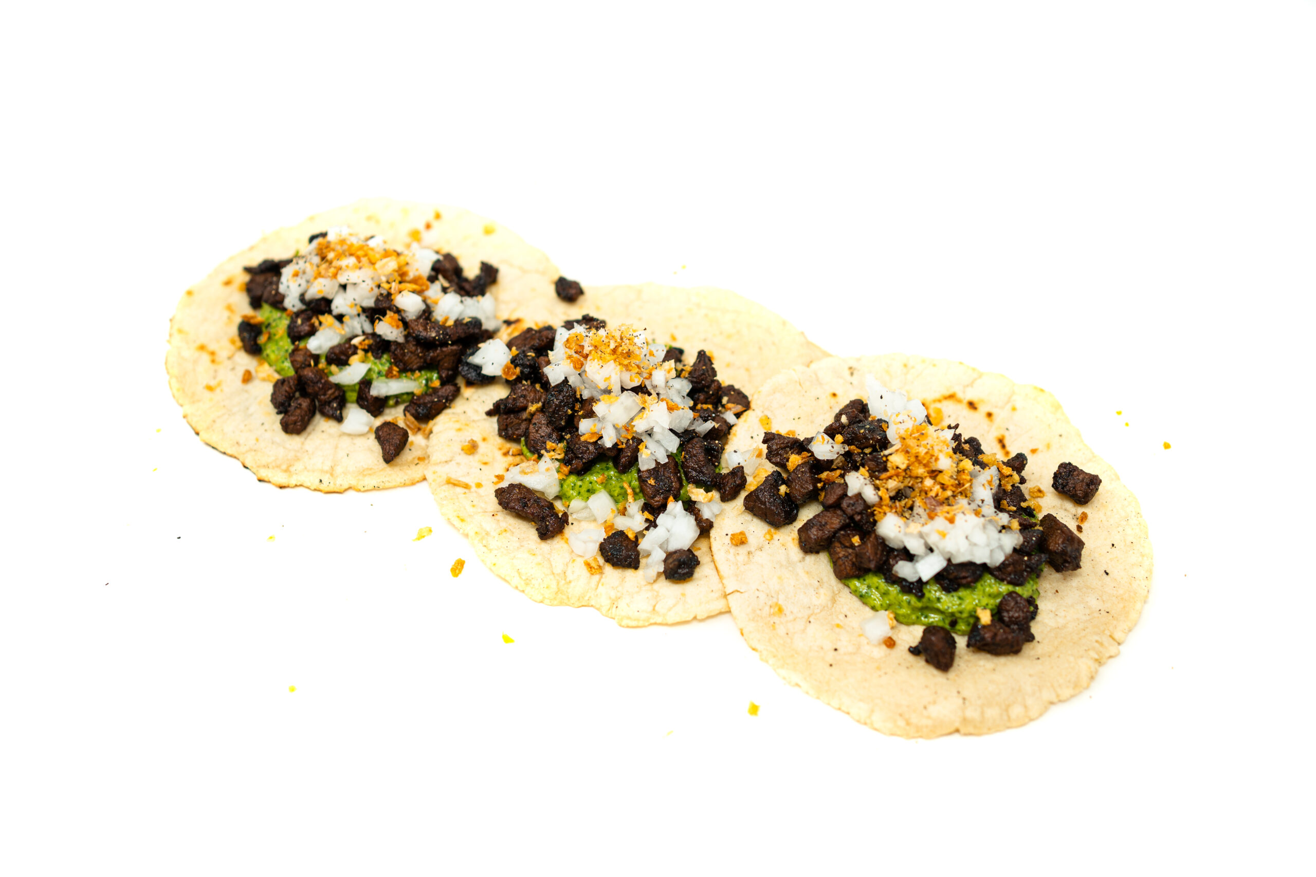 Close up on toasted corn tortillas loaded with glossy, dark-seared cubes of glazed steak, sitting on a thick base of vibrant green chimichurri under warm directional light.
