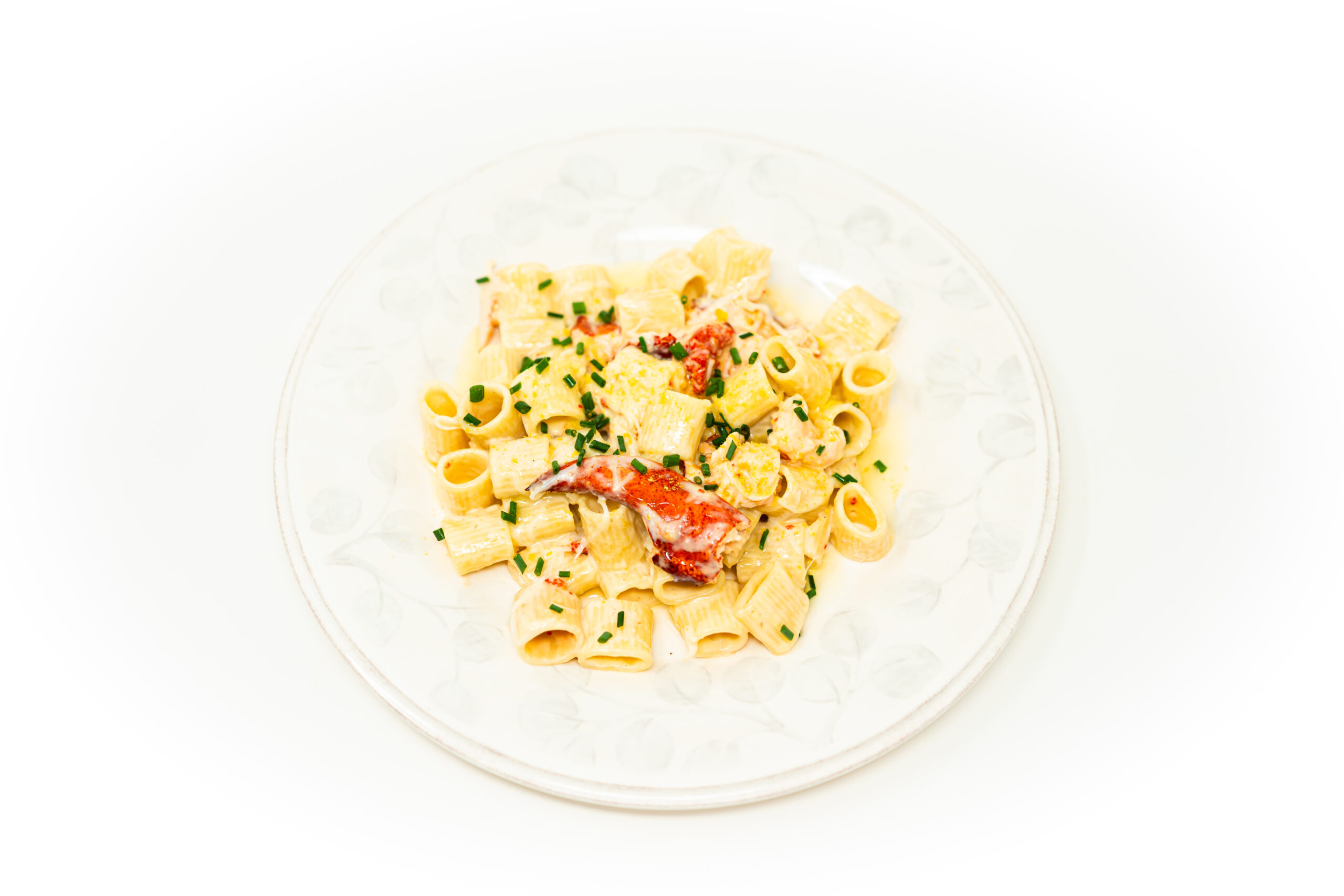 Top-down shot of mini rigatoni in a glossy cheese sauce topped with lump crab, a butter-glazed lobster claw, chives, and a blackened charred lemon half on dark slate.
