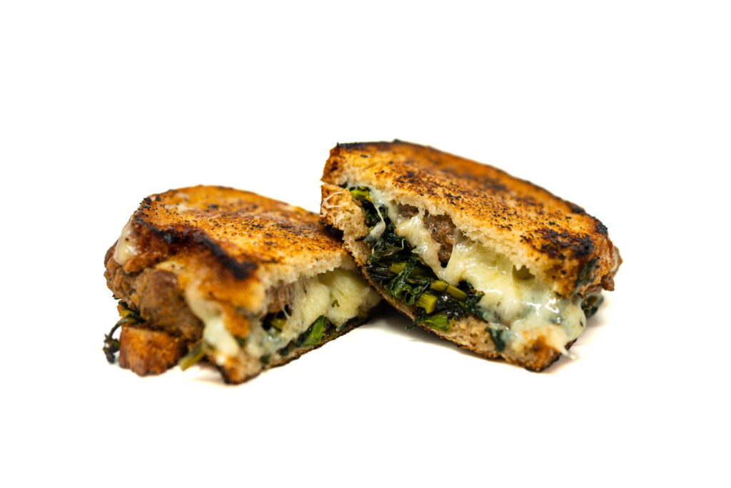 A cross-section of a toasted sourdough sandwich revealing a thick, juicy lamb patty, melted Gruyère cheese, and dark green charred rapini, resting on a dark slate board.