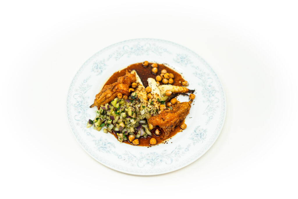 Close-up angled shot of pan-fried flatfish split to reveal its white steamed interior, surrounded by dark brown agrodolce sauce, raw purple pepper brunoise, fried squash blossoms, and blistered chickpeas under bright, hard lighting.