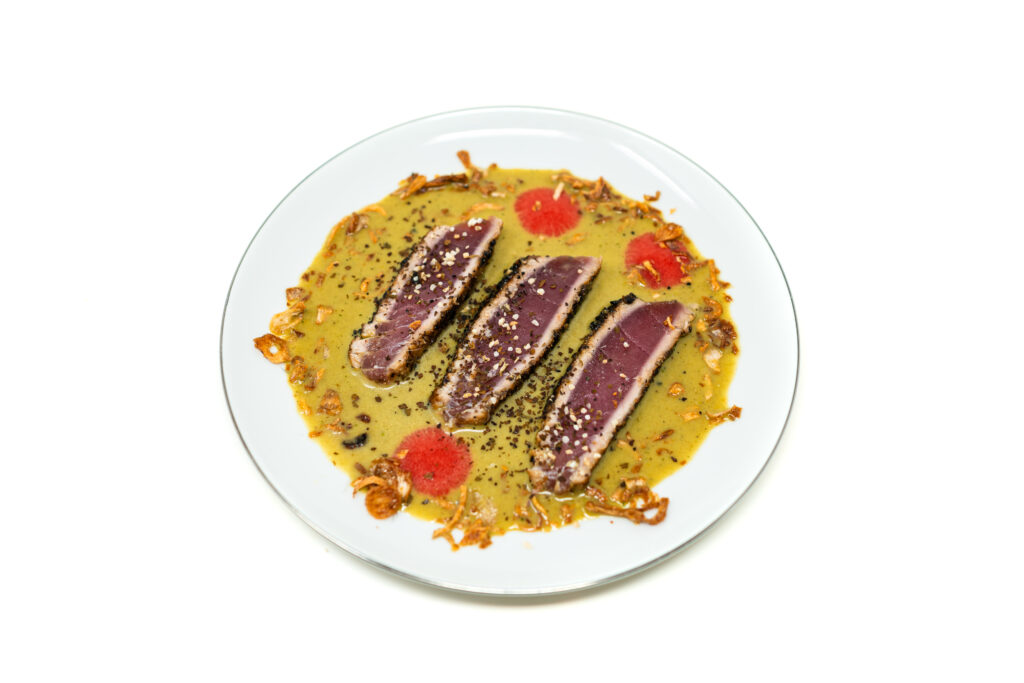 Overhead view under bright, flat lighting showing three slices of rare ahi tuna with a dark, textured spice crust. The fish is shingled in the center of a white plate over a shallow pool of opaque green broth, surrounded by three glossy red dots of fluid gel and scattered brittle fried shallots.