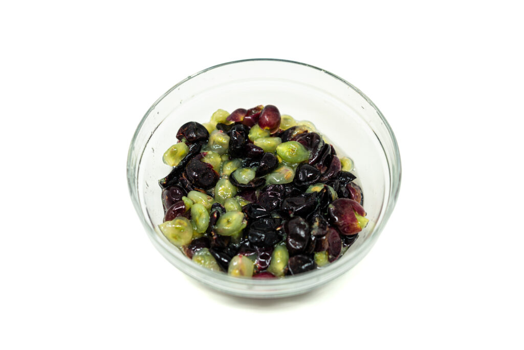A clear glass bowl centered on a stark white background containing a wet, glossy mixture of torn, dark purple muscadine hulls and pale, translucent interior grape flesh under bright overhead lighting.