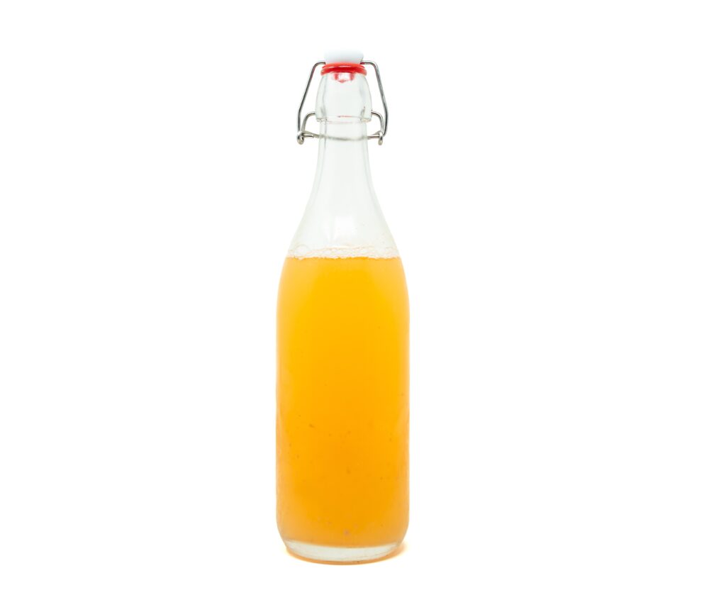 A swing-top glass bottle filled with a bright, opaque golden liquid, showing tight carbonation bubbles at the surface against a stark white background.
