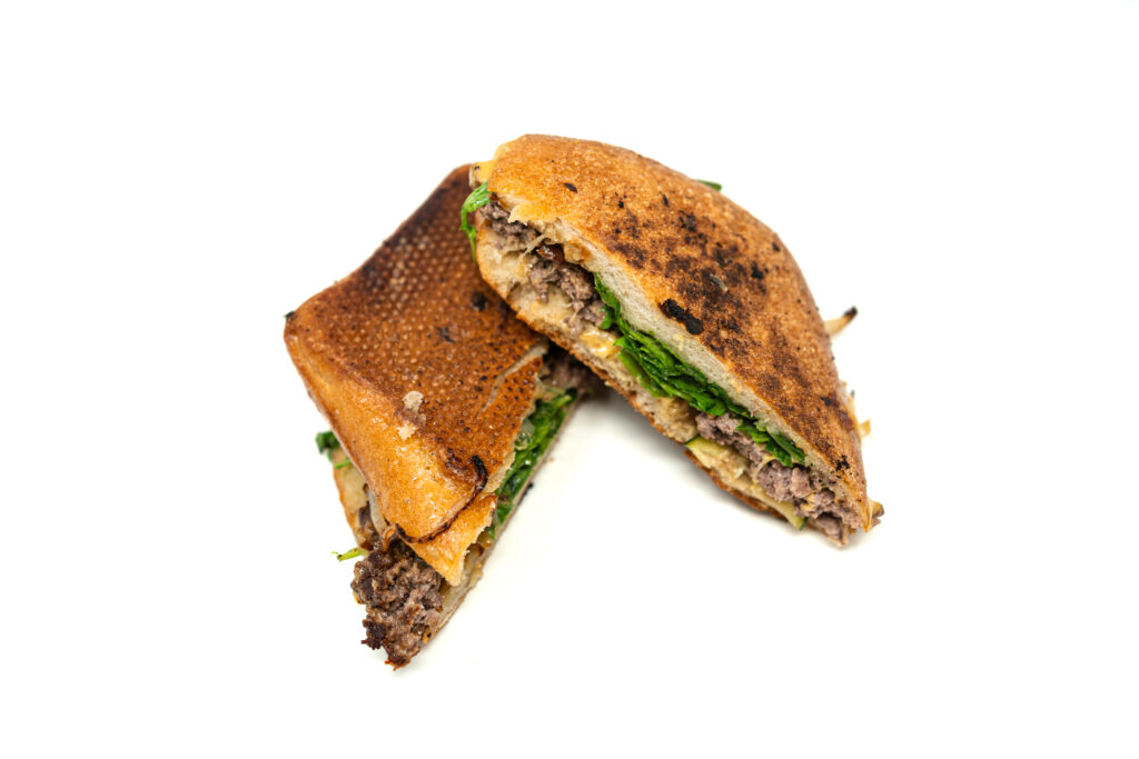 An overhead perspective of a halved ciabatta smash burger isolated on a white surface. The direct lighting emphasizes the uneven, porous texture and dark, charred high points of the pressed ciabatta crust. The cut edges showcase the compressed physical layers of the sandwich, with the lacy edges of the ground beef and wilted arugula extending just past the perimeter of the bread.