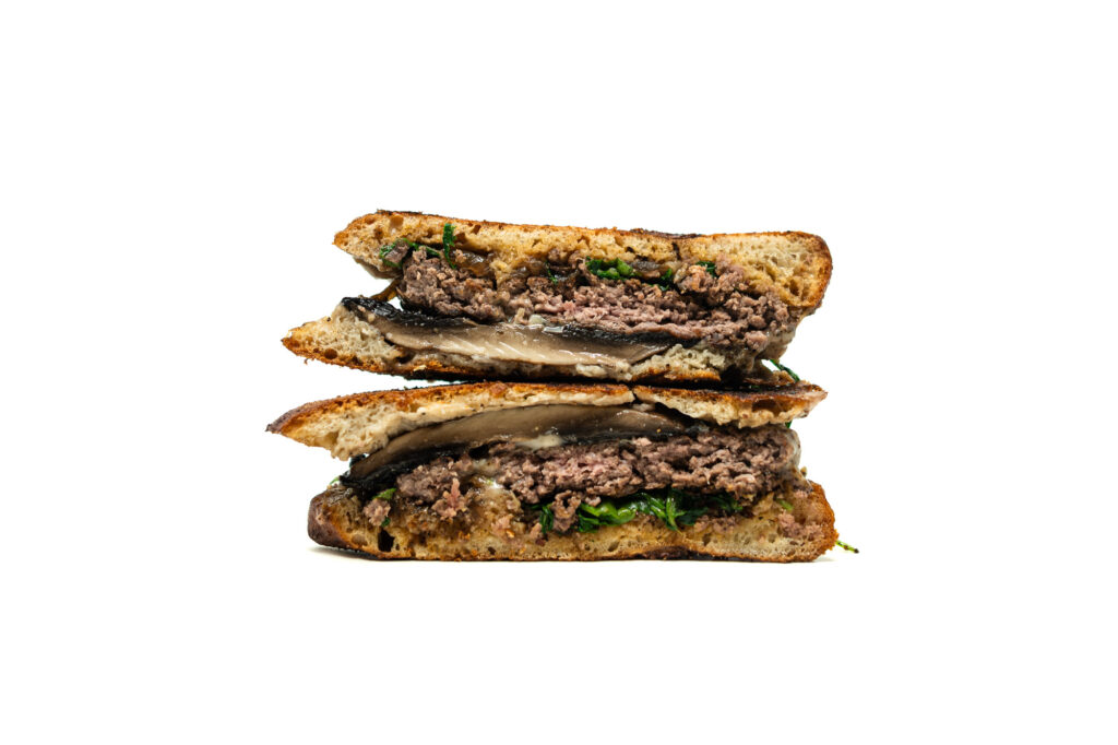 A tight, vertically stacked cross-section of a halved burger against a white background. The profile view emphasizes the physical textures of the build, showing a coarse beef patty, a thick portobello mushroom cap, wilted arugula, glossy melted white cheese, and dark bacon fig jam compressed between deeply browned ciabatta
