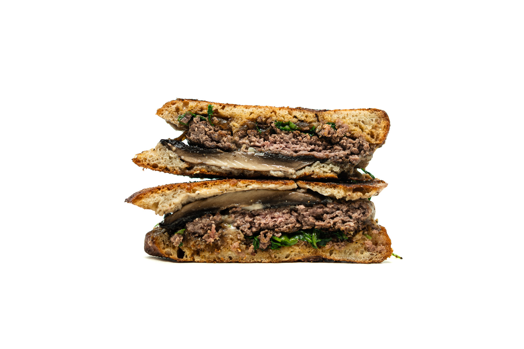 A tight, vertically stacked cross-section of a halved burger against a white background. The profile view emphasizes the physical textures of the build, showing a coarse beef patty, a thick portobello mushroom cap, wilted arugula, glossy melted white cheese, and dark bacon fig jam compressed between deeply browned ciabatta