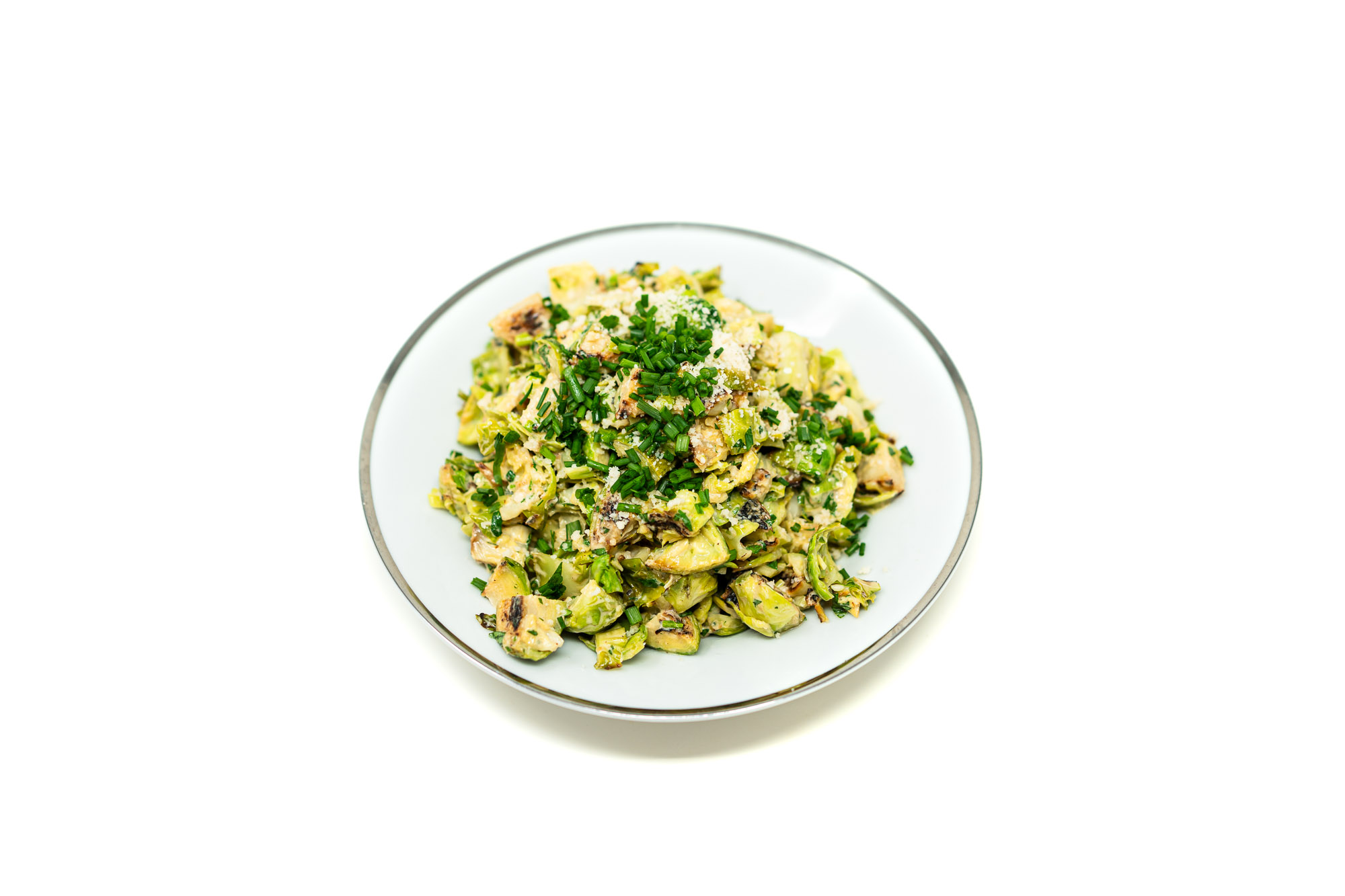 High-angle overhead shot of Caesar Dressed Broiled Nukazuke Brussels Sprouts served on a thin-rimmed white plate. The quartered sprouts display dark charred edges coated in a creamy dressing, topped with a dense mound of finely minced green chives and a dusting of grated Parmigiano-Reggiano.