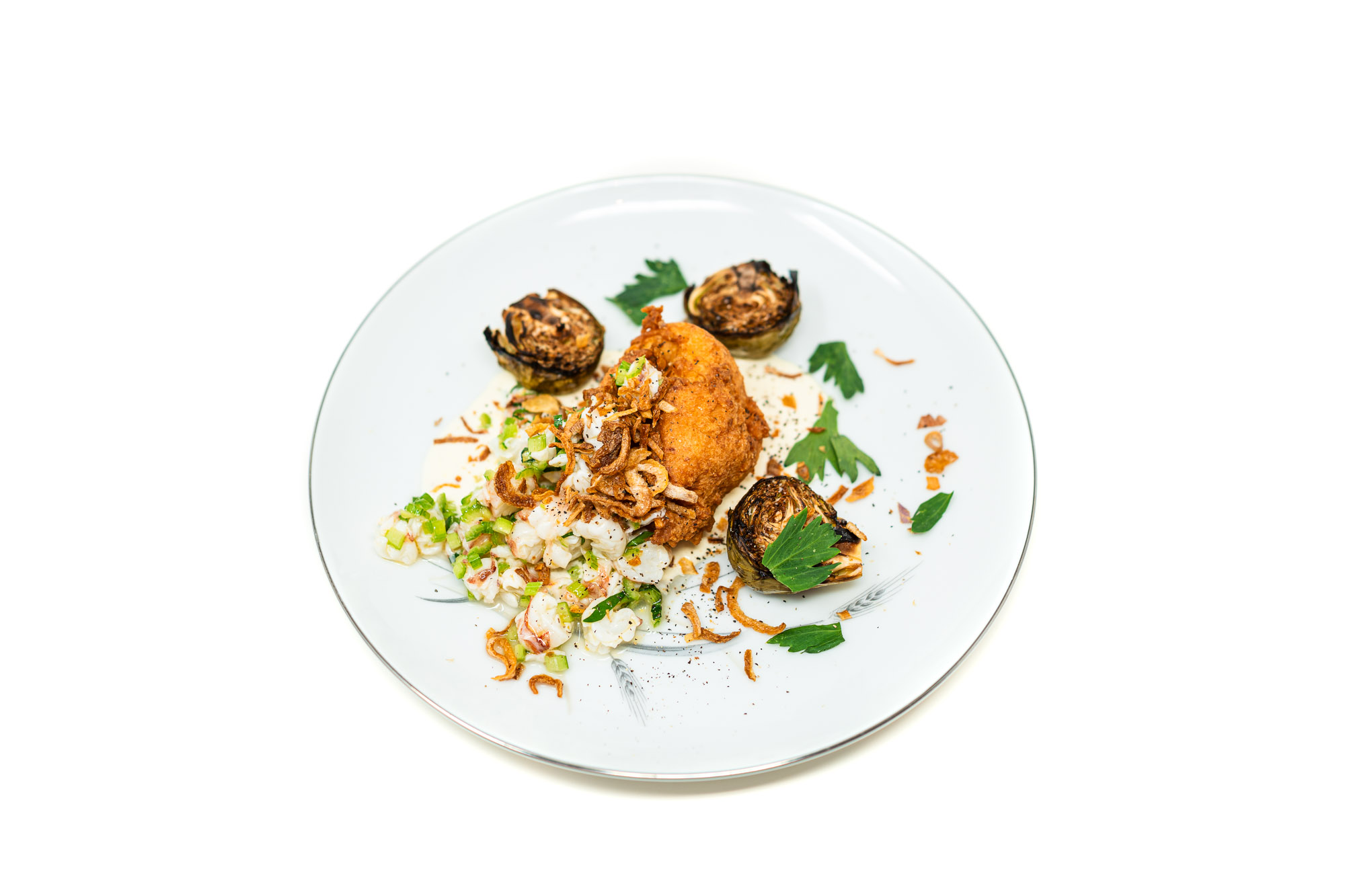 A deeply crusted, golden shrimp fritter rests in the center of a creamy emulsion next to a bright, finely diced shrimp tartare, surrounded by three heavily charred Brussels sprout halves under stark, directional lighting.